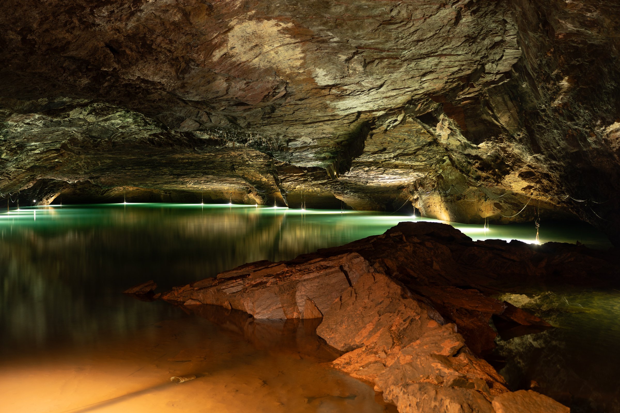 Underground cave with glowing green and amber lake reflected off stalactite ceiling